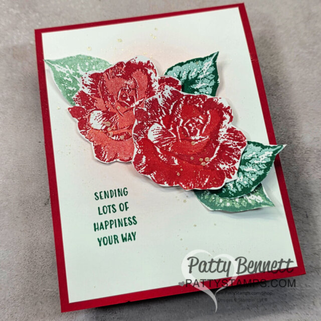 Handmade card with two large red and white roses and green leaves. Text below reads, “Sending LOTS OF HAPPINESS YOUR WAY.” Background is white with gold splatter, red border, and watermark “Patty Bennett pattystamps.com” at lower right. To read more, visit Patty Stamps (pattystamps.com)