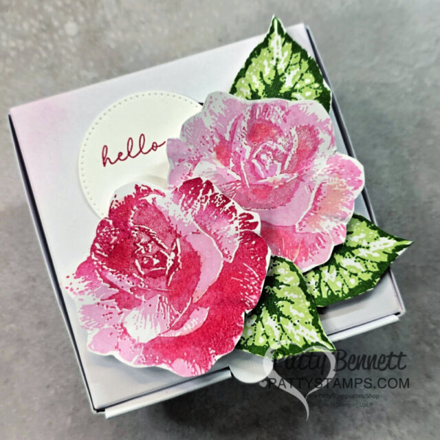 A small white gift box is decorated with two realistic pink and red stamped roses, two green stamped leaves, and a white circular tag with "hello" written in pink cursive. The box rests on a gray surface. “PATTY BENNETT” watermark is in the lower right. To read more, visit Patty Stamps (pattystamps.com)