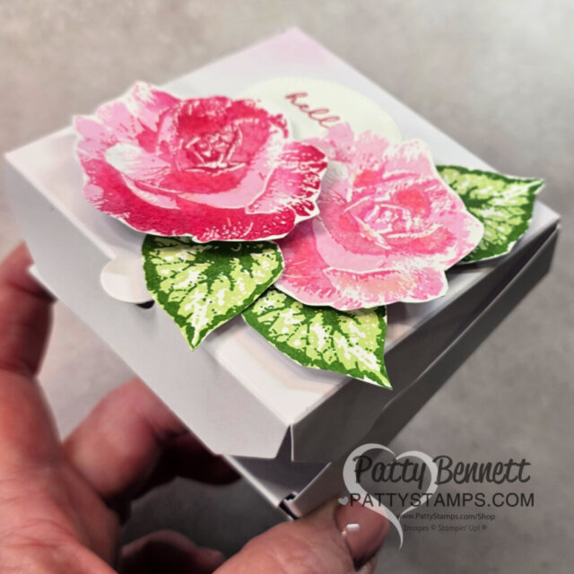 A hand holds a small white gift box decorated with two pink paper roses and green leaves. The box lid features the word "hello" in light pink and is partially open. The image is watermarked with "Patty Bennett PattyStamps.com. To read more, visit Patty Stamps (pattystamps.com)
