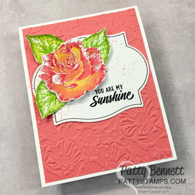 Handmade greeting card with embossed coral background, a white label with "You Are My Sunshine" in black text, and a vibrant orange-yellow rose with green leaves. "Patty Bennett pattystamps.com" watermark is on the lower right corner. To read more, visit Patty Stamps (pattystamps.com)