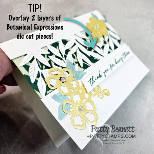 A hand holds a handmade card decorated with layered white and colored floral and leaf die cuts. Text reads “TIP! Overlay 2 layers of Botanical Expressions die cut pieces!” and “thank you for being there.” The card features yellow and blue flowers with gem accents. To read more, visit Patty Stamps (pattystamps.com)
