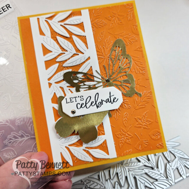 A handmade greeting card features a bright orange embossed background with a leafy pattern, a vertical white die-cut leaf border, and a gold metallic butterfly. A white tag with “LET’S celebrate” in black script is attached. The card sits on a clear stamp sheet. To read more, visit Patty Stamps (pattystamps.com)