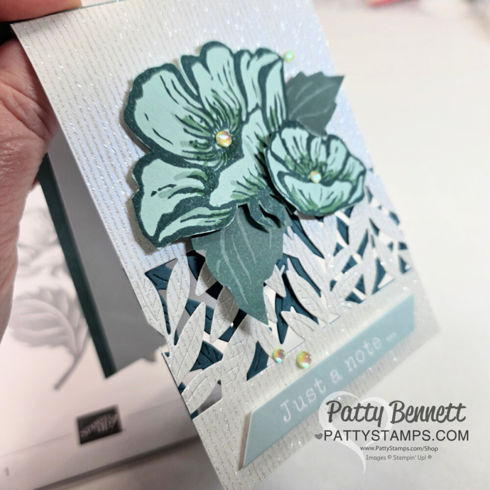 A hand holds a handcrafted greeting card decorated with layered green floral die-cuts, rhinestones, and a white lattice design. The card features the text "Just a note..." on a light blue banner. The background has a textured white pattern. To read more, visit Patty Stamps (pattystamps.com)