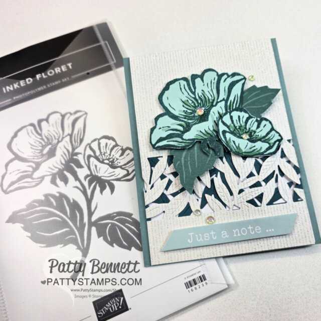 A handmade card featuring layered, teal flowers with green leaves over a white woven pattern. A label at the bottom reads “Just a note …” The card sits beside a floral stamp set labeled “Inked Floret.” The artist’s name, Patty Bennett, appears below. To read more, visit Patty Stamps (pattystamps.com)