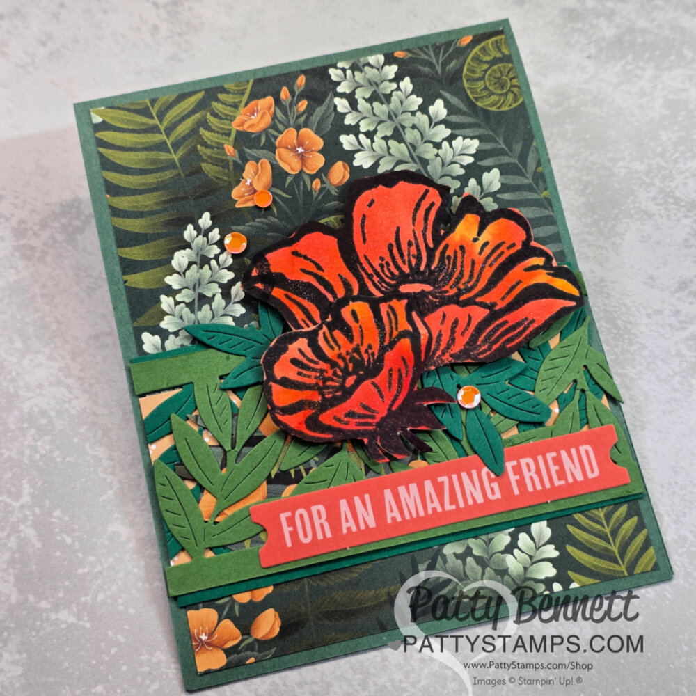 A handmade card featuring bold red-orange flowers, green leaves, small orange blossoms, and gems. The background is dark green with a floral pattern. A coral banner reads “For an Amazing Friend.” The card is signed "Patty Bennett, PattyStamps.com.". To read more, visit Patty Stamps (pattystamps.com)