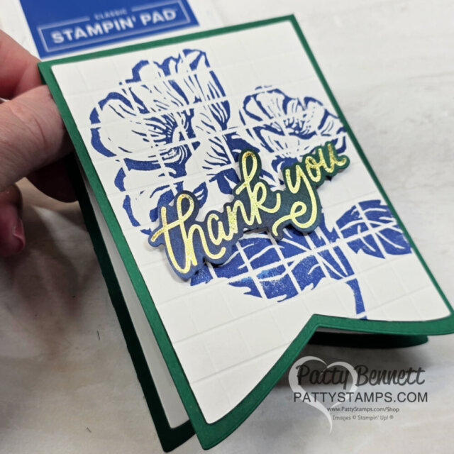 A hand holds a green-bordered card with embossed white, featuring blue floral art and a gold “thank you” on top. A blue SU! ink pad and “PattyStamps.com” watermark are visible. The card has a pointed edge at the bottom. To read more, visit Patty Stamps (pattystamps.com)