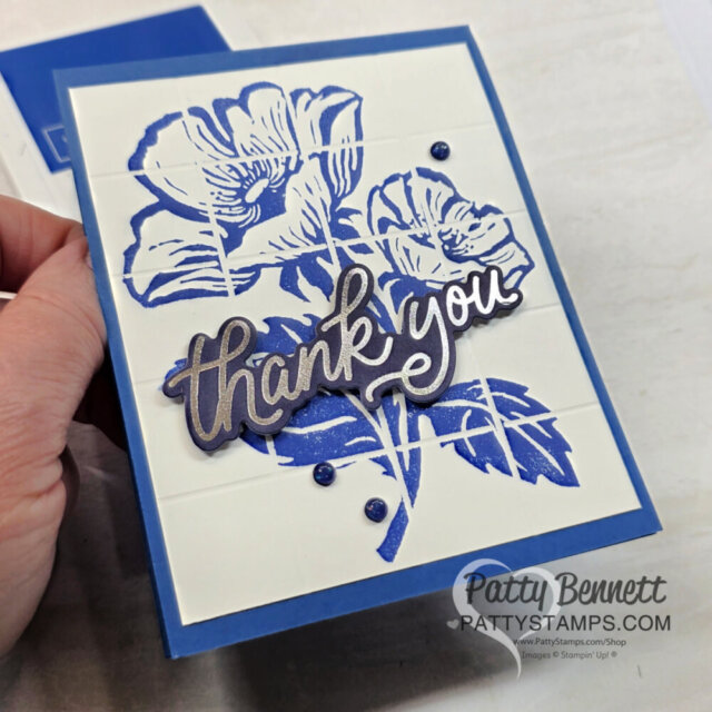 A hand holds a blue and white floral card with raised squares, a large blue flower design, a metallic “thank you” cutout, and small blue pearls. “Patty Bennett PattyStamps.com” is watermarked in the corner. To read more, visit Patty Stamps (pattystamps.com)