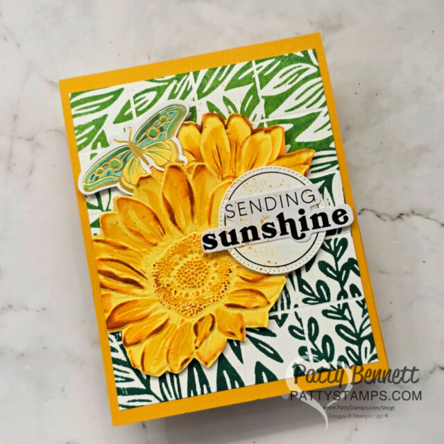 A handmade card features a vibrant yellow sunflower, a green and yellow butterfly, and a "Sending sunshine" greeting on a white background with green leaf patterns. The card is displayed on a light marbled surface. To read more, visit Patty Stamps (pattystamps.com)