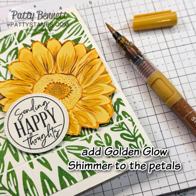 A handmade card with a large yellow sunflower on green leaf background, a "Sending Happy Thoughts" circle tag, and a yellow shimmer pen nearby. Text reads: "add Golden Glow Shimmer to the petals." Grid paper underneath. To read more, visit Patty Stamps (pattystamps.com)