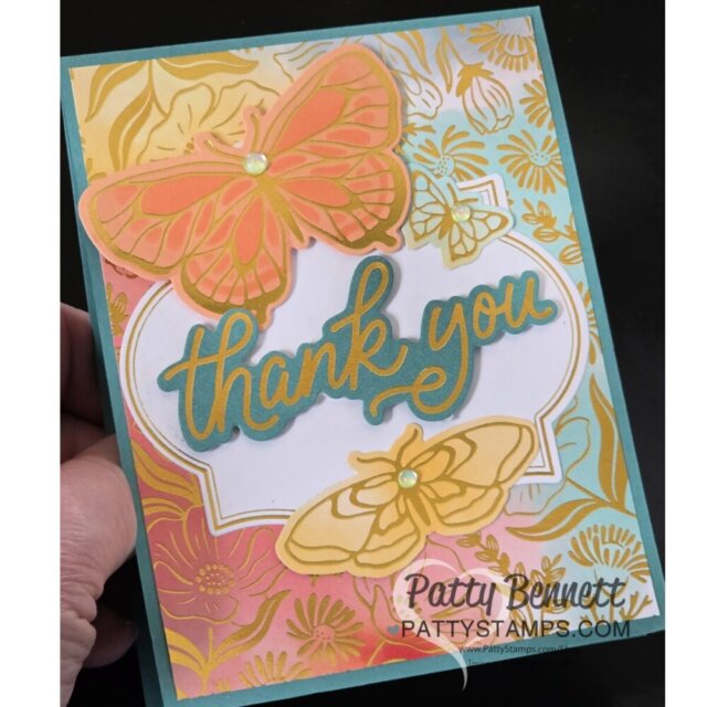 Hand holding a colorful greeting card with layered orange and yellow butterflies, gold floral accents, and “thank you” in bold blue and gold script at the center. The bottom right corner reads “Patty Bennett Pattystamps.com.”. To read more, visit Patty Stamps (pattystamps.com)