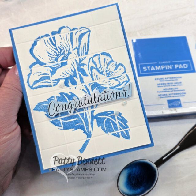 A hand holds a handmade card featuring a large blue floral design and the word “Congratulations!” in script. The card has a tiled background. Nearby are a blue ink pad labeled “Stampin’ Pad” and a blending brush with blue ink. To read more, visit Patty Stamps (pattystamps.com)