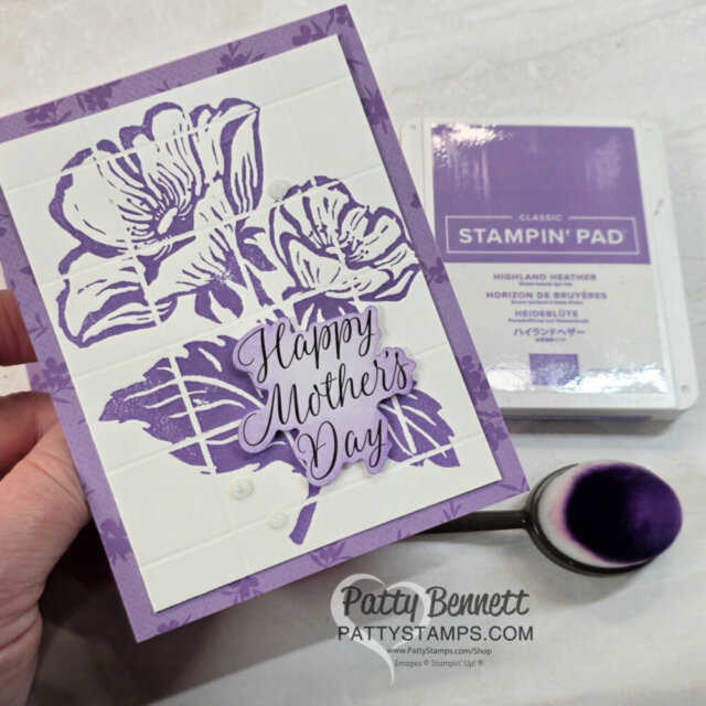 A hand holds a purple floral "Happy Mother’s Day" card made with stamped iris flowers and leaves. Next to it are a Highland Heather Stampin’ Pad, an ink blending brush with purple bristles, and a mat labeled “Patty Bennett pattystamps.com.”. To read more, visit Patty Stamps (pattystamps.com)