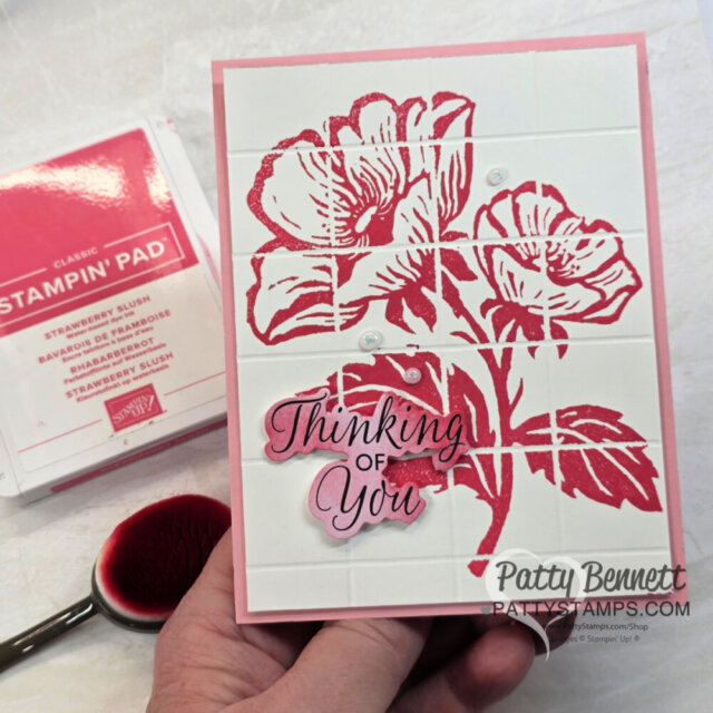 A hand holds a handmade card featuring pink stamped flowers and leaves on a white grid-patterned background. The card has a raised "Thinking of You" sentiment and pearl embellishments. A pink ink pad and blending brush are visible in the background. To read more, visit Patty Stamps (pattystamps.com)