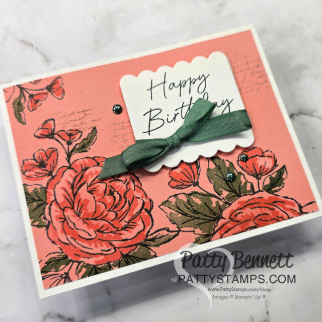 A floral card with pink roses and green leaves on a coral background. A scalloped white label reads “Happy Birthday” in script. A green bow and several green pearls decorate the card. “Patty Bennett” and her website watermark appear at the bottom. To read more, visit Patty Stamps (pattystamps.com)