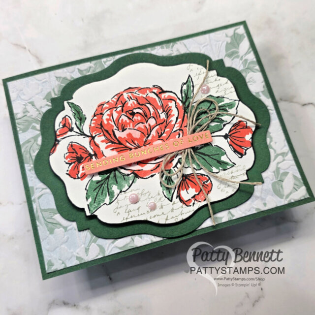 Handmade card featuring a large red and pink rose with green leaves, layered on ornate green and white frames. A gold-embossed pink strip reads “Sending bunches of love.” Twine bow and soft floral background. “Patty Bennett” watermark at bottom right. To read more, visit Patty Stamps (pattystamps.com)