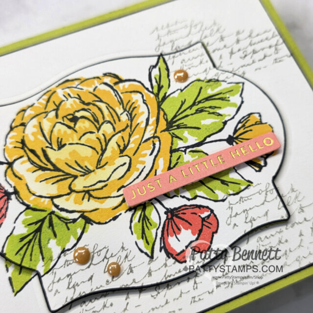 A handmade card features a large yellow and orange flower with green leaves, accented by two coral buds. Script handwriting forms the background. A pink banner with gold text reads "JUST A LITTLE HELLO." Small brown embellishments decorate the card. To read more, visit Patty Stamps (pattystamps.com)