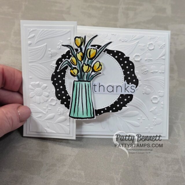 A handmade card with embossed white floral patterns, a black-and-white polka-dot circle frame, and a turquoise vase holding yellow tulips. The word "thanks" is written across the center. A hand holds the card, which is partially open. To read more, visit Patty Stamps (pattystamps.com)