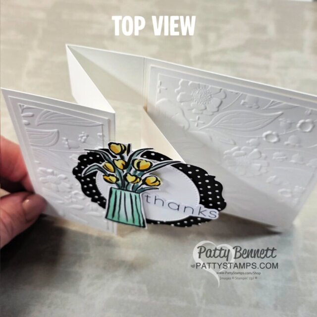 A hand holds a white embossed greeting card with a zigzag fold. The card features a polka-dot oval, yellow flowers in a green vase, and text reading "thanks." "Top View" is written at the top. The watermark says "PATTYSTAMPS.COM Patty Bennett. To read more, visit Patty Stamps (pattystamps.com)