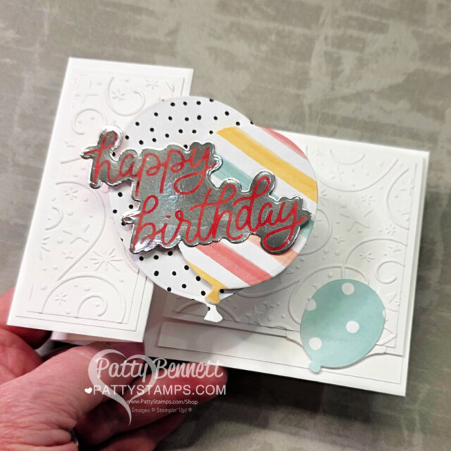 A hand holds a white, textured pop-up birthday card with embossed swirls, a pastel striped balloon, and red glitter "happy birthday" text. There’s also a blue polka-dot balloon. The card has layered, dimensional elements and bold, festive design. To read more, visit Patty Stamps (pattystamps.com)