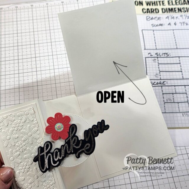A handmade thank you card partly open, showing embossed white floral patterns, a red flower, and bold black "thank you" text. An arrow points to the unfolded panel labeled "OPEN." A craft mat and instruction sheet are visible in the background. To read more, visit Patty Stamps (pattystamps.com)