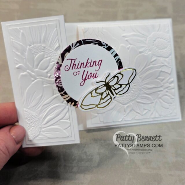 A hand holds a white embossed floral card with “Thinking of You” in purple text. The message is on a round centerpiece, accented by a metallic border and gold butterfly. The background features raised sunflower patterns. "Patty Bennett pattystamps.com" is watermarked. To read more, visit Patty Stamps (pattystamps.com)