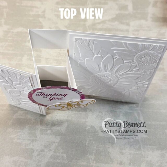 Top view of a handmade white card with embossed sunflower designs and a pop-out panel. The front panel has a circular tag reading "Thinking of You" with a yellow bow. Background is a light, slightly textured surface. “Patty Bennett” watermark is visible. To read more, visit Patty Stamps (pattystamps.com)